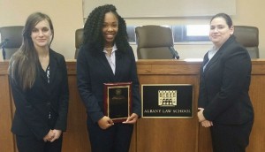 Three women wearing suits pose for a photo with the woman in the center holding a plaque