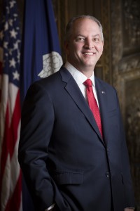 A man wearing a navy blue suit and red tie smiles with the flags of Louisiana and the United States in the background