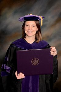 A female student wearing a graduation cap and gown and holding a diploma smiles
