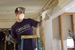 A man wearing a dust mask and goggle rips off dry wall from a house