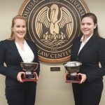 Two female students wearing black suits and white shirts and holding silver trophies for a photo with the LSU Law Center seal in the background