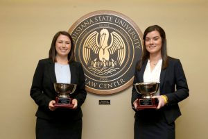 Two female students wearing black suits and white shirts and holding silver trophies for a photo with the LSU Law Center seal in the background