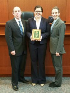 Three students pose for a photo with the student in the middle holding a plaque