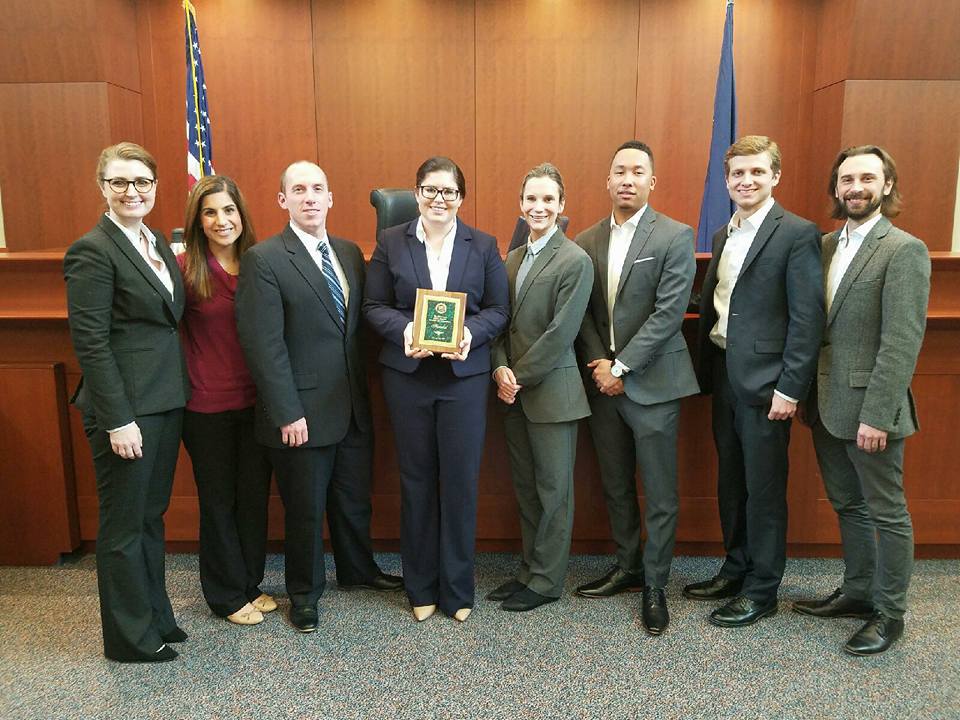 Eight students wearing suits pose for a photo with one student holding a plaque and a courtroom in the background