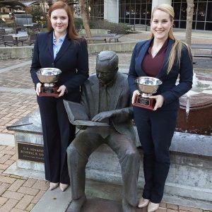 Two female students wearing suits hold silver trophies and pose next to a statue outside the LSU Law Center plaza