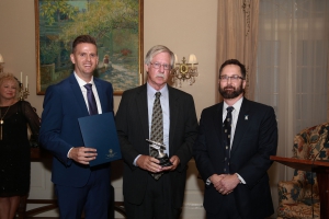 Three men wearing suits pose for a photo with two of them holding awards and a painting in the background