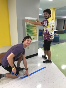 Two male students hang signs and put tape on the floor