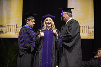 female graduate smiles while two male faculty members place hood on her