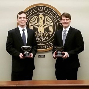 Two male students wearing suits hold silver trophies and stand in front of the LSU Law Center seal