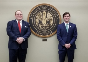 Two male students wearing suits pose for a photo next to the LSU Law logo