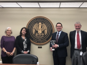 A male student holds a crystal trophy and stands next to a man and two women next to the LSU Law Center seal