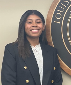 Britney Young wearing a black blazer with gold buttons and a white blouse smiling in front of beige wall and the LSU Law seal.