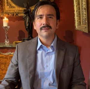 Alvaro de la Cruz-Correa in a gray blazer and blue button down shirt sitting in front of a table with a lit candle and statue. 