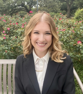 Kayla Meyers wearing a gray blazer with a white button down smiling and sitting on a bench in front of flowers and bushes. 