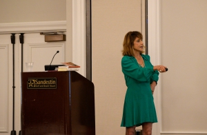 Professor Marlene Krousel in a green dress pointing her clicker to the right as she presents in front of an audience.